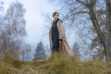 A beautiful girl with long hair with wireless headphones in her ears in a raincoat walks on the grass in a deserted place against the background of autumn trees and looks back