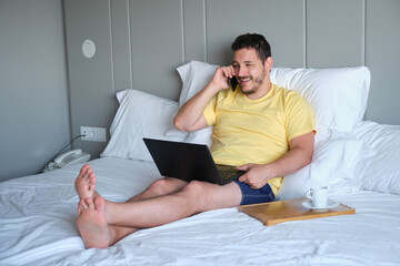 Desperate man speaks on the phone and works with laptop in an hotel room bed.