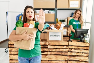 Young brunette woman at wearing volunteer t shirt holding bag with food with open hand doing stop sign with serious and confident expression, defense gesture