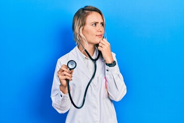 Beautiful young blonde doctor woman holding stethoscope with hand on chin thinking about question, pensive expression. smiling with thoughtful face. doubt concept.