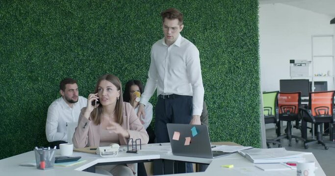 Manager boss having an argument with employee in office, scolds for talking on phone at work place. Female office worker in formal wear sit talking on phone with someone, distracted from work