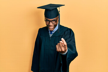 Young african american man wearing graduation cap and ceremony robe beckoning come here gesture with hand inviting welcoming happy and smiling