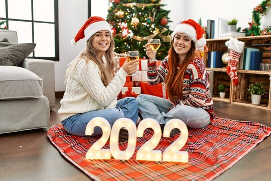 Woman couple toasting with champagne for new year 2022 sitting by christmas tree at home