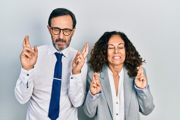 Middle age couple of hispanic woman and man wearing business office uniform gesturing finger crossed smiling with hope and eyes closed. luck and superstitious concept.