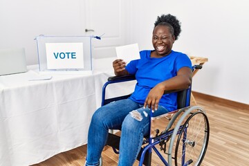 Young african woman sitting on wheelchair voting putting envelop in ballot box winking looking at...