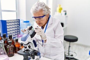 Senior grey-haired woman wearing scientist uniform using microscope at laboratory