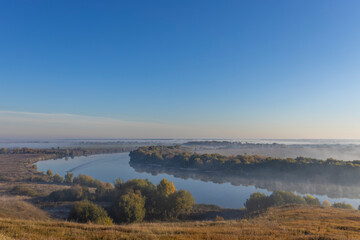 Autumn landscape in the early morning overlooking the river. A wide river and endless expanses of fields. Yellow leaves on trees and bushes are illuminated by the rays of the rising sun.