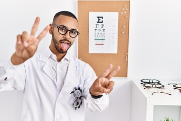 African american optician man standing by eyesight test smiling with tongue out showing fingers of both hands doing victory sign. number two.