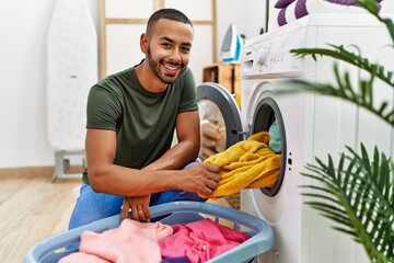 Young hispanic man cleaning clothes using washing machine at laundry - obrazy, fototapety, plakaty