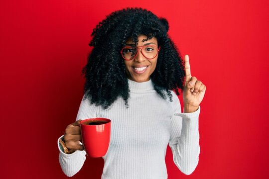 African american woman with afro hair holding coffee smiling with an idea or question pointing finger with happy face, number one - Powered by Adobe