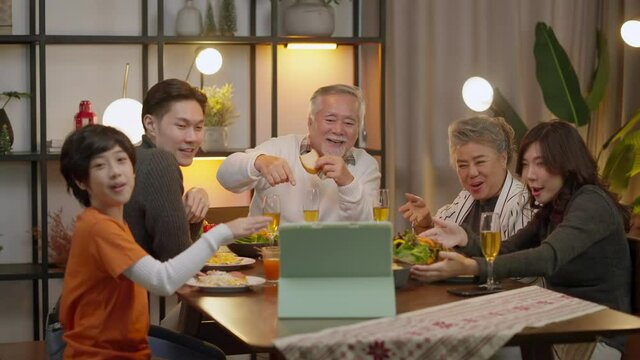 happiness asian multi generation have dinner together while video calling conference to distance relative friend using tablet device wireless internet connection,family waving hand to tablet say hi