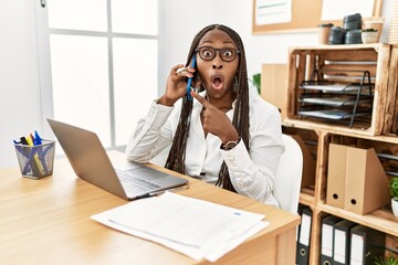 Black woman with braids working at the office speaking on the phone surprised pointing with finger to the side, open mouth amazed expression.