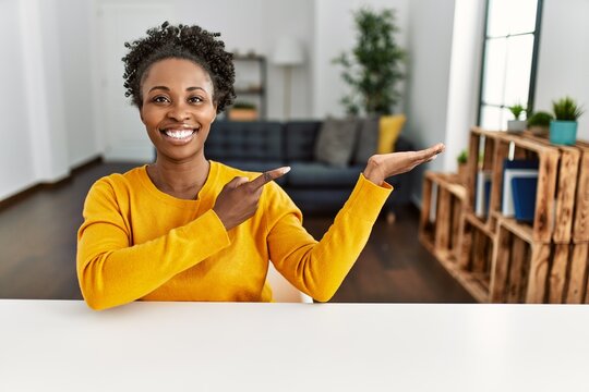 Young african american woman wearing casual clothes sitting on the table at home amazed and smiling to the camera while presenting with hand and pointing with finger.