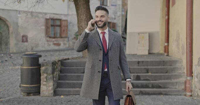 bearded young man in suit with long coat holding suitcase, talking on the phone and walking in front of medieval city