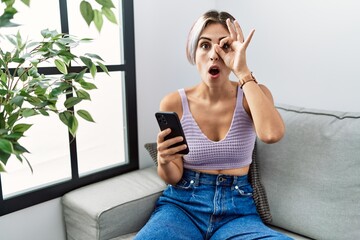 Young beautiful woman using smartphone typing message sitting on the sofa doing ok gesture shocked with surprised face, eye looking through fingers. unbelieving expression.