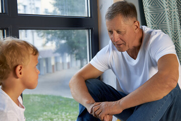 Modern grandfather sitting at the window transmits life experience to his grandson. Child listens...