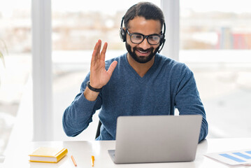 Friendly Indian male employee wearing headset and eyeglasses making online consultation, holding...