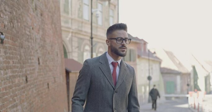 sexy young businessman wearing suit and long coat, fixing eyeglasses, looking to side and walking away 