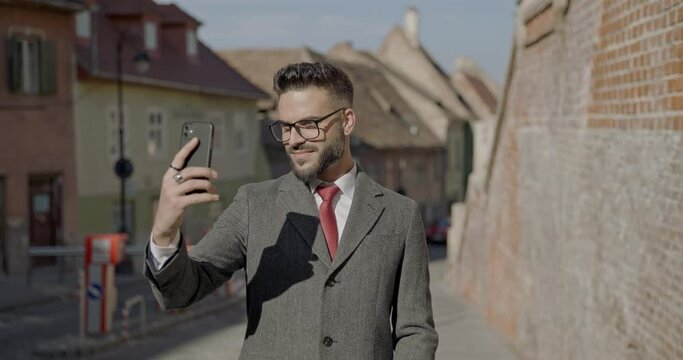 handsome young businessman in elegant suit holding phone and taking selfies, smiling outside in front of medieval town