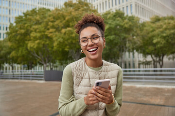Outdoor shot of curly haired woman wears casual jumper and vest uses mobile phone reads news from...