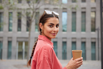 Sideways shot of pretty woman with pigtail dressed in casual red shirt holds paper cup of coffee enjoys aromatic drink while strolling in city poses against blurred background. Lifestyle concept