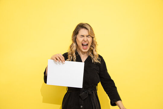 young beautiful woman loudly shouts, negative emotions, holds white blank sheet of paper in hands A4, copy space for inscription, studio shot on yellow background
