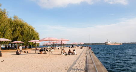 Toronto, Canada - 08 29 2021: Torontonians in downtown Toronto having rest under pink umblrellas of the Sugar Beach bestde blue waters of lake Ontario with a cargo ship