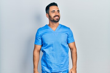 Handsome hispanic man with beard wearing blue male nurse uniform looking away to side with smile on...