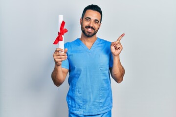 Handsome hispanic man with beard wearing blue male nurse uniform holding diploma smiling happy...