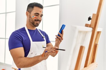 Young hispanic man smiling confident drawing using smartphone at art studio