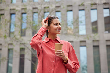 Positive young female tourist wears spectacles on head and red shirt holds disposable paper cup drinks aromatic coffee looks away with happy expression poses against blurred building background