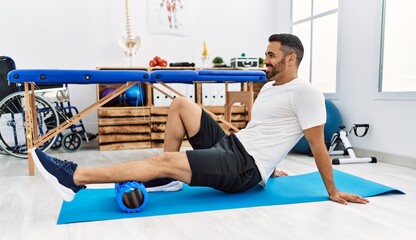 Young hispanic man patient smiling confident having leg rehab session using foam roller at clinic