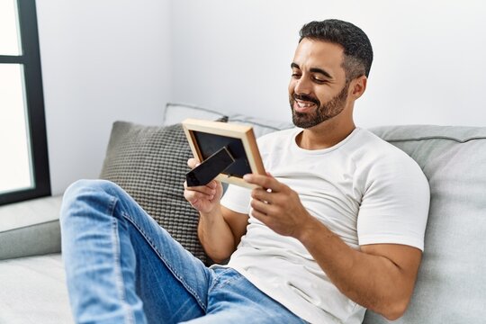 Young hispanic man smiling confident looking photo frame at home