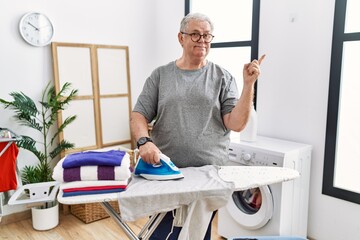 Obraz premium Senior caucasian man ironing clothes at home smiling happy pointing with hand and finger to the side