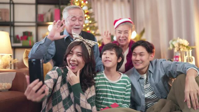 Smiling asian big family with little child boy kids sit on couch in living room make self-portrait video on cell together, happy parents with small children have fun videocall on smartphone at home 