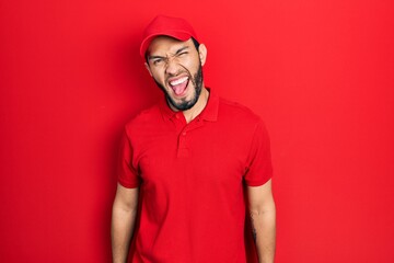 Hispanic man with beard wearing delivery uniform and cap sticking tongue out happy with funny expression. emotion concept.