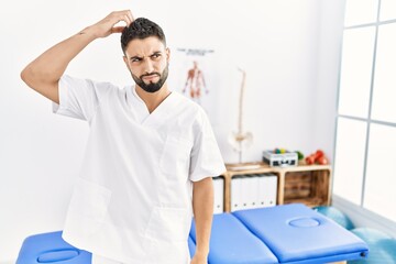 Young handsome man with beard working at pain recovery clinic confuse and wonder about question. uncertain with doubt, thinking with hand on head. pensive concept.