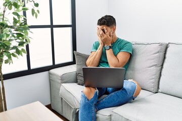 Young handsome man with beard using computer laptop sitting on the sofa at home with sad expression covering face with hands while crying. depression concept.