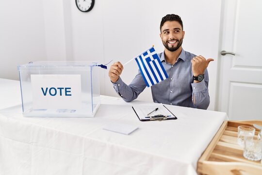 Young handsome man with beard at political campaign election holding greece flag pointing thumb up to the side smiling happy with open mouth