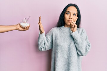 Young hispanic girl rejecting bowl with sugar serious face thinking about question with hand on...