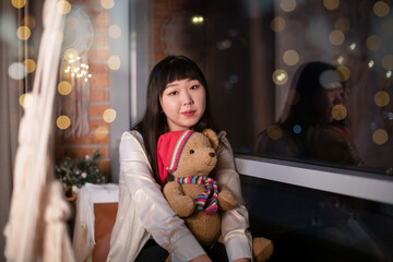 A cute lonely young Korean Asian woman holds a funny soft bear in her arms, sitting on the window at the night window of the house. The concept of New Year and Christmas