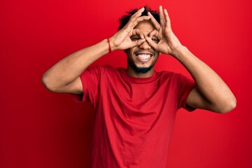 Young african american man with beard wearing casual red t shirt doing ok gesture like binoculars sticking tongue out, eyes looking through fingers. crazy expression.