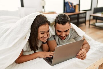Young latin couple watching movie covering with bed sheet on the bed.