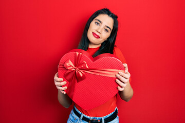 Young hispanic girl holding valentine gift relaxed with serious expression on face. simple and natural looking at the camera.