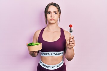 Young hispanic girl wearing sportswear eating salad puffing cheeks with funny face. mouth inflated...