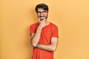 Young hispanic man wearing casual clothes and glasses looking confident at the camera smiling with crossed arms and hand raised on chin. thinking positive.