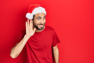 Young hispanic man wearing christmas hat smiling with hand over ear listening an hearing to rumor or gossip. deafness concept.