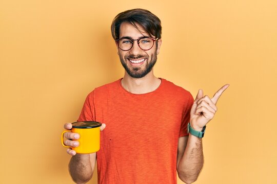 Young hispanic man drinking a cup of coffee smiling happy pointing with hand and finger to the side