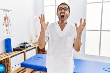 Obraz premium Middle age man with beard working at pain recovery clinic crazy and mad shouting and yelling with aggressive expression and arms raised. frustration concept.