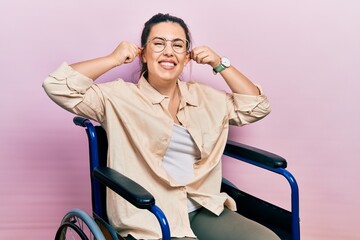 Young hispanic woman sitting on wheelchair smiling pulling ears with fingers, funny gesture. audition problem